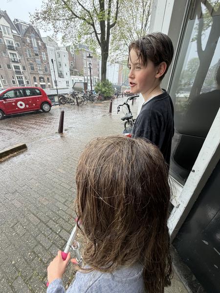 Two children stand on a wet, cobblestone street in Amsterdam, specifically Oostersekade, Uilenburg, with classic canal houses lining the background